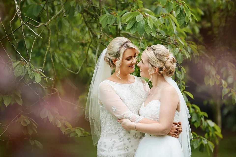 Two brides smiling together beneath green branches